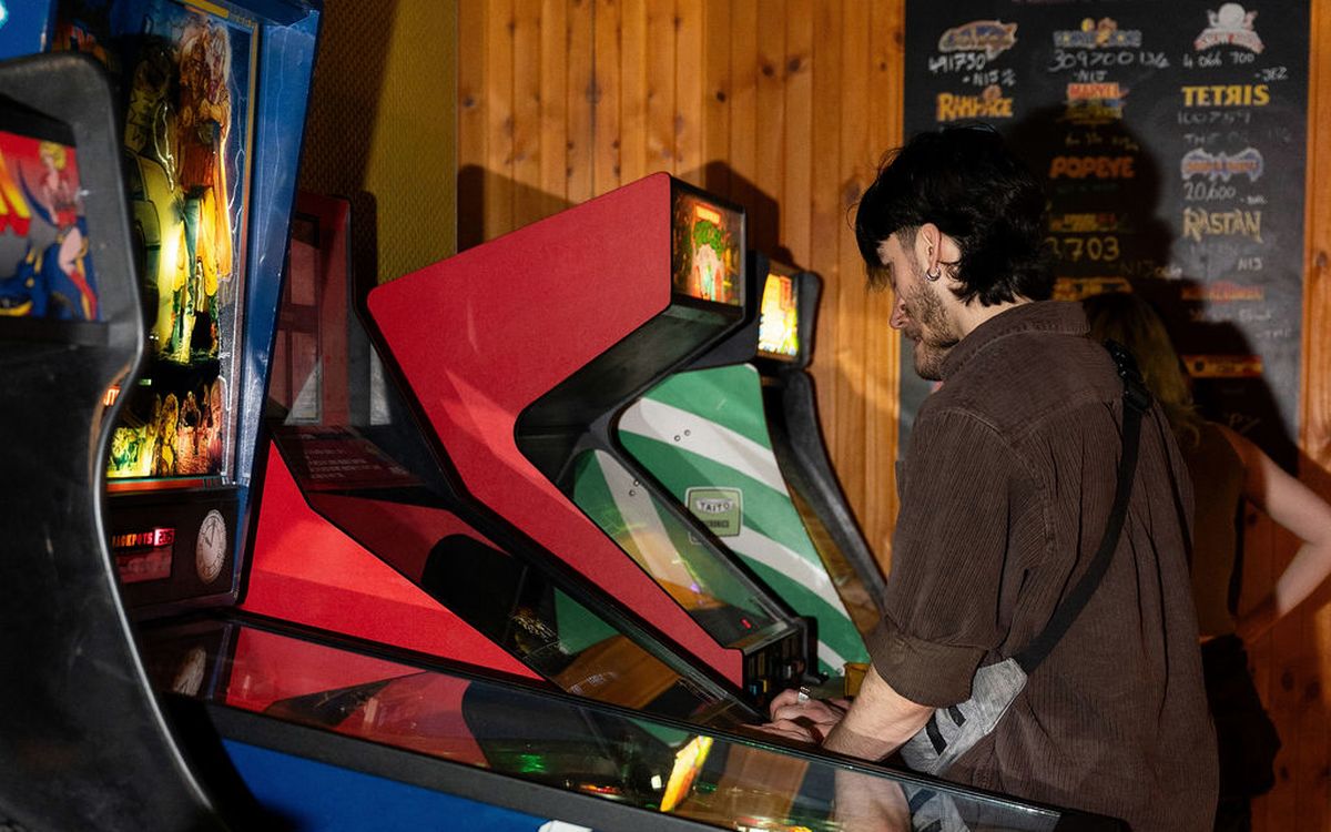 A guest playing pinball beside the arcade cabinets, high score board chalked up behind
